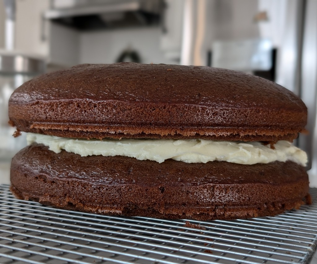 Chocolate cake with cream filling rests on a cooling rack in a kitchen. The fluffy texture and rich brown color stand out.