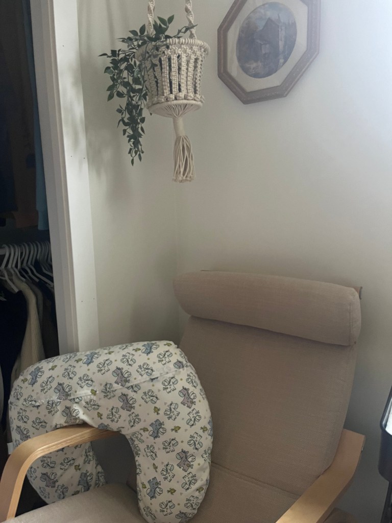 Beige chair with patterned nursing pillow, hanging plant in macramé holder, and framed picture on white wall, creating a cozy, serene space.