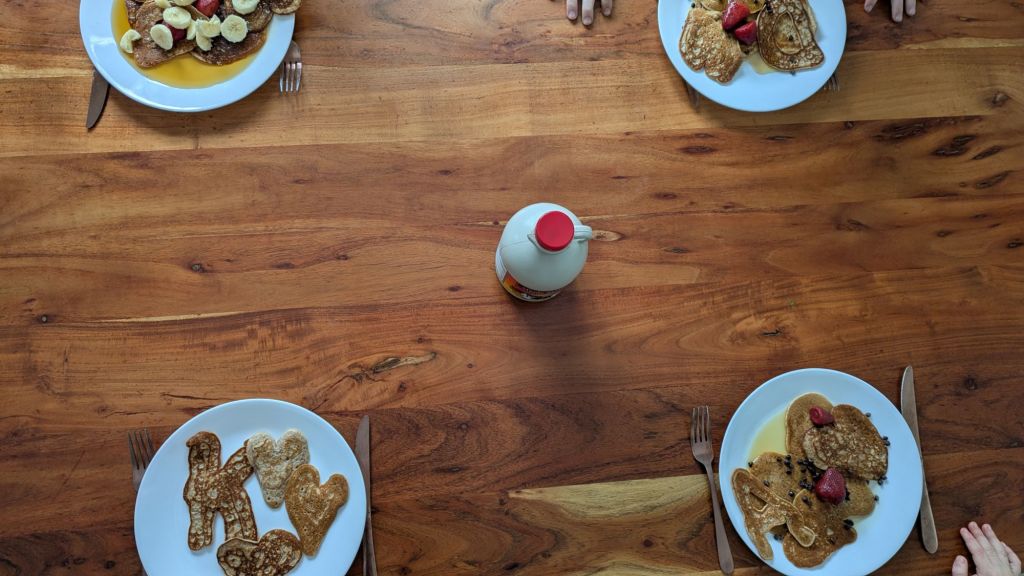 Plates of pancakes with fruit and syrup on a wooden table, hands visible, bottle of syrup in the center. Warm, inviting breakfast scene.