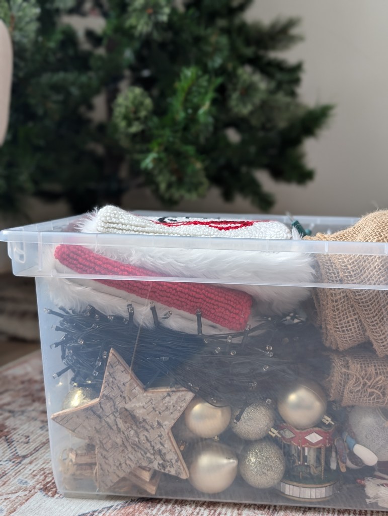 Plastic bin filled with Christmas decorations like stockings, lights, and ornaments. A decorated tree is in the blurred background.