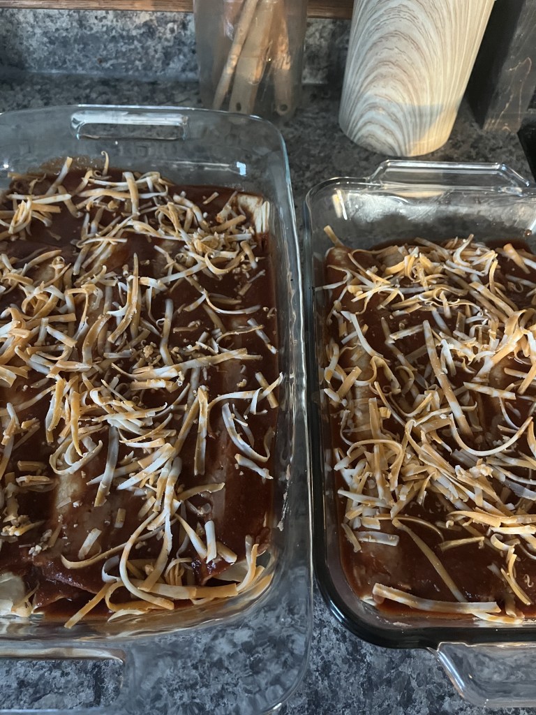 Two glass baking dishes filled with enchiladas topped with red sauce and shredded cheese on a gray countertop. Cooking utensils in the background.