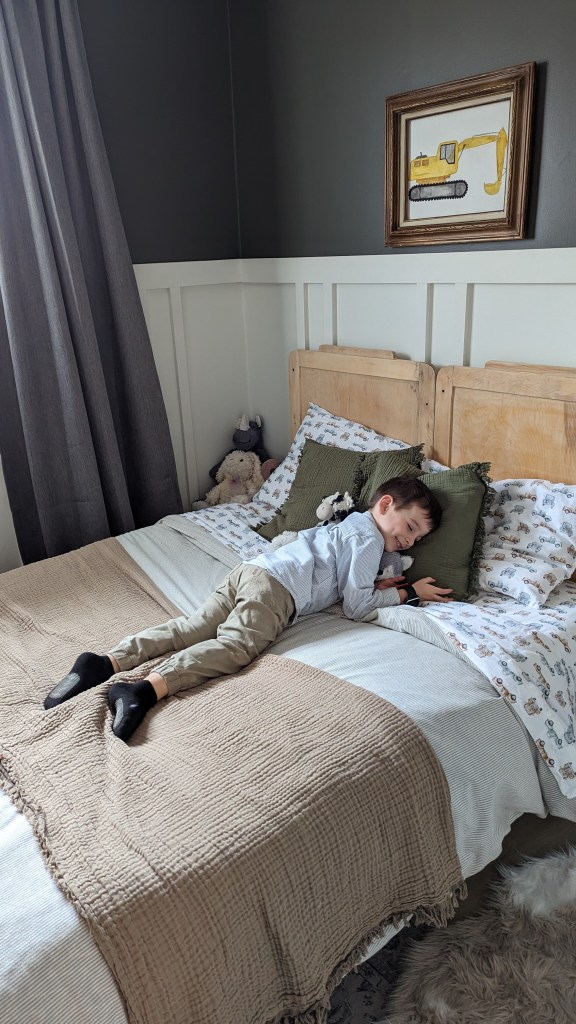 Child laying on a bed with a beige blanket, green pillow, and stuffed toy. Dark curtains and a framed yellow excavator drawing on the wall.