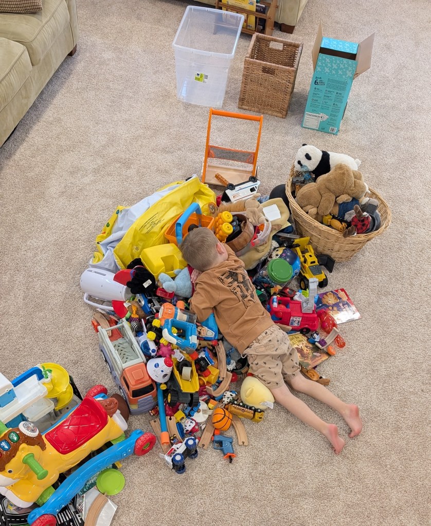 Child in brown shirt lying on a pile of colorful toys on a carpeted floor, surrounded by baskets, creating a playful, relaxed scene.