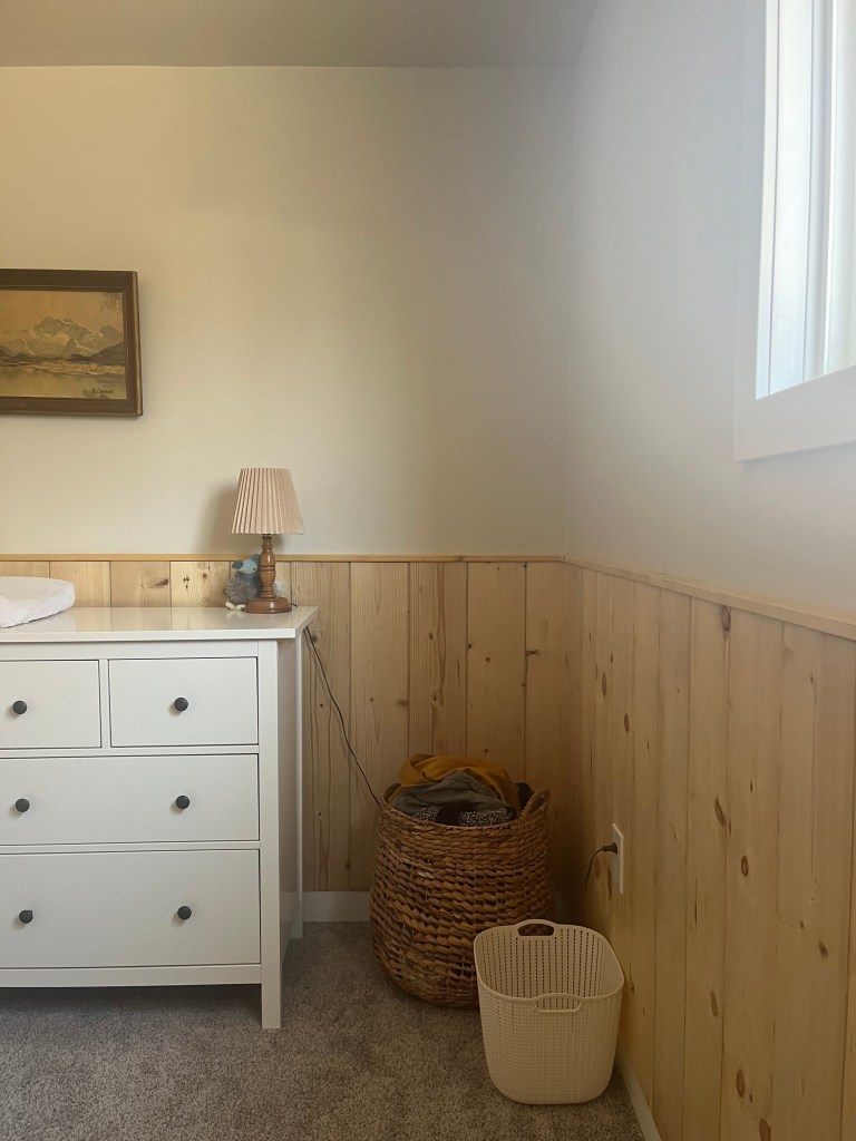 White dresser with lamp and toy, wicker and white baskets on gray carpet, wood-paneled walls, painting above, peaceful room.