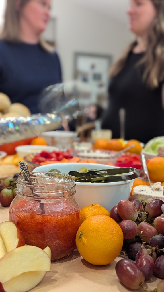Two people talking in a blurred background, with a foreground of a charcuterie of diverse foods, a jar of jam, and a cheerful tabletop setting.