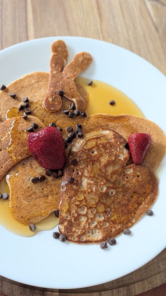Pancakes in fun shapes with chocolate chips, strawberries, and syrup on a white plate. Wooden table in the background.