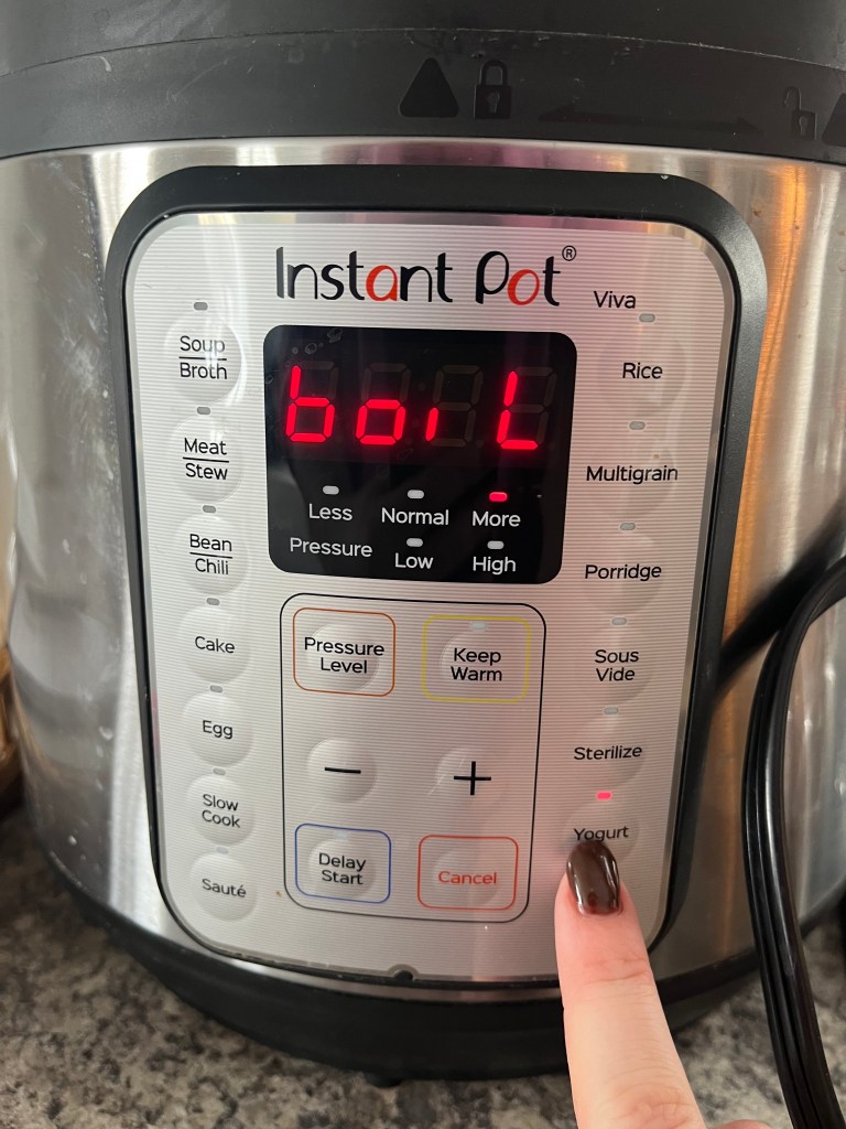 Finger pressing "Yogurt" button on Instant Pot with display showing "boil." Controls in black and red; stainless steel background.