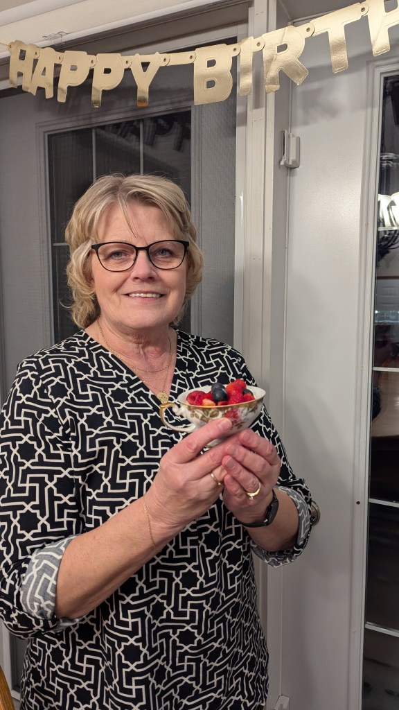 Smiling woman holding dessert with berries. "Happy Birthday" banner in background. Wearing black-and-white patterned top.