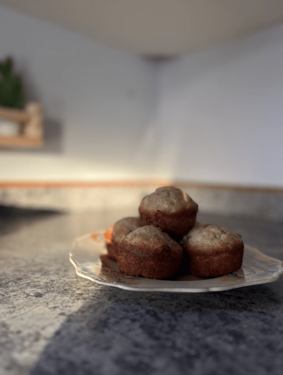 Plate of four muffins on a gray countertop, warmly lit. Background is blurred, with a hint of green, creating a cozy kitchen atmosphere.