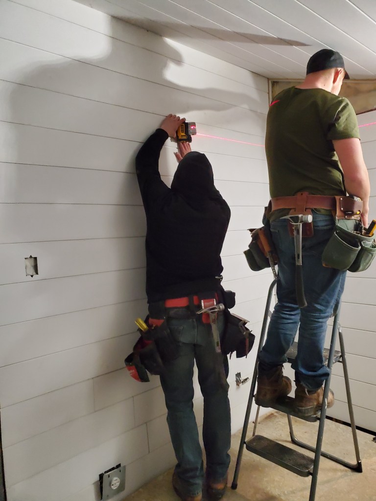Two people installing white paneling in a room. One uses a drill while the other stands on a ladder. Tools are visible in tool belts.