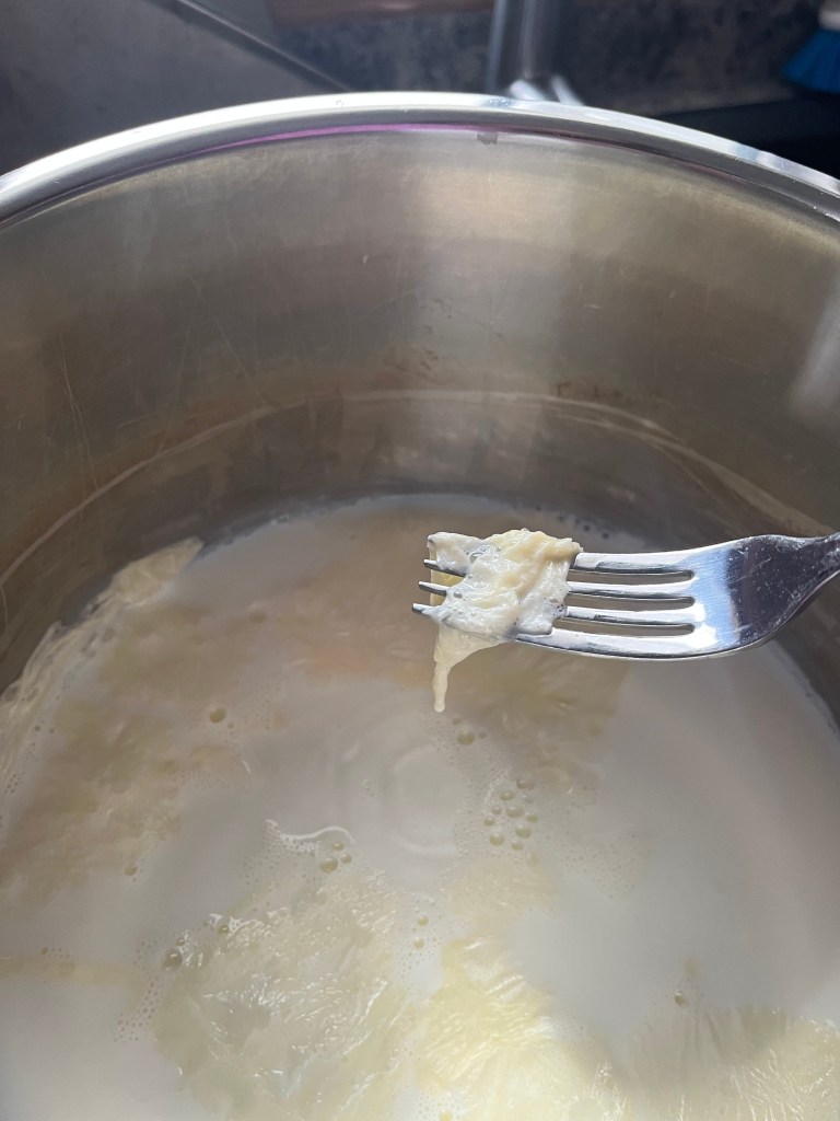 Fork holding a cheese curd over a pot of milk, set in a kitchen. The scene is calm, with soft light reflecting off the metal pot.