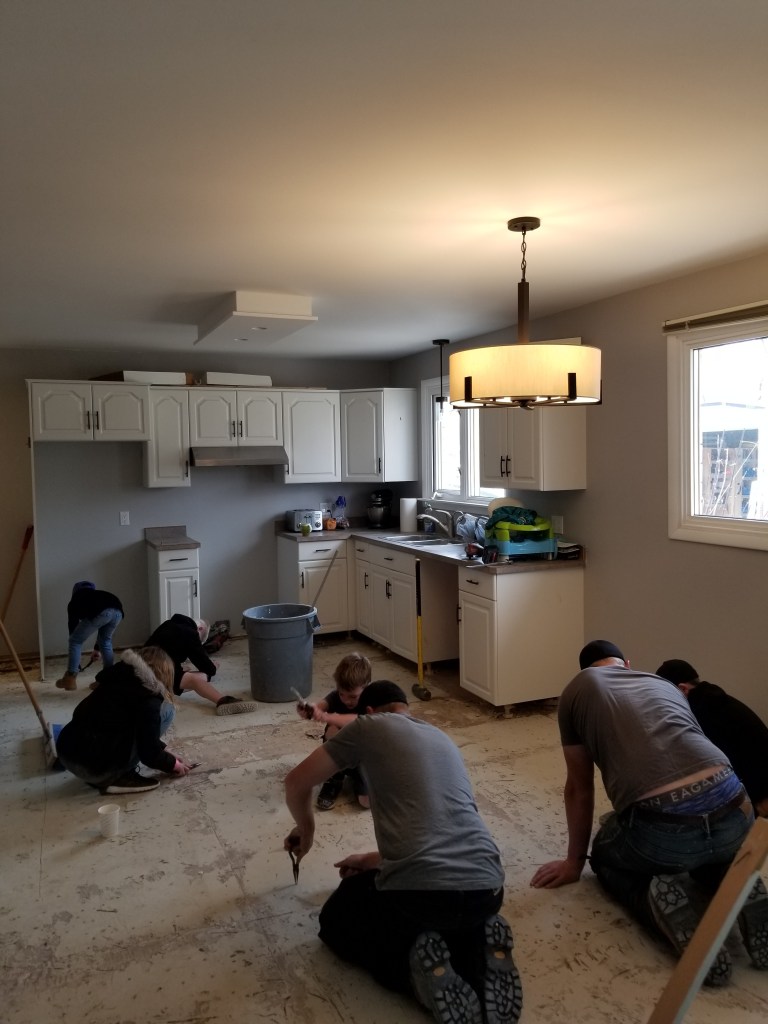 Five people working on a kitchen floor renovation, kneeling and using tools. White cabinets and a large pendant light are visible.