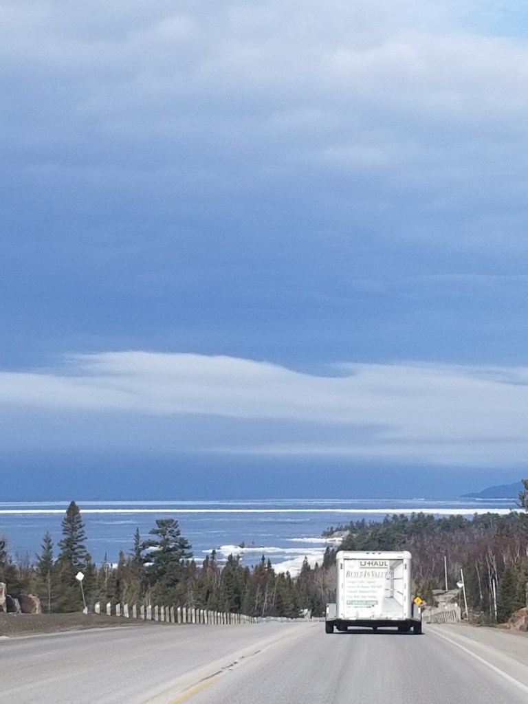 A U-Haul trailer travels on a road beside trees and a frozen lake under a cloudy blue sky, evoking a calm, cold atmosphere.