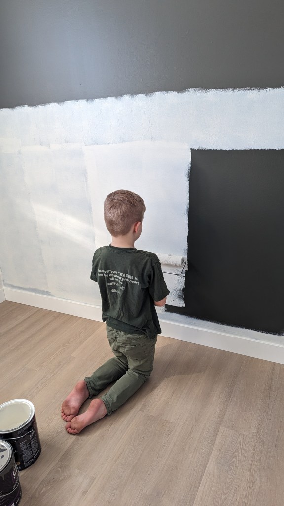 A child kneels, painting a dark wall white, in a room with wooden flooring. Paint cans are nearby. The mood is focused and creative.