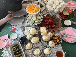 People seated around a table with assorted pastries, sandwiches, and fruits on a green tablecloth. Plates and pink napkins are set.
