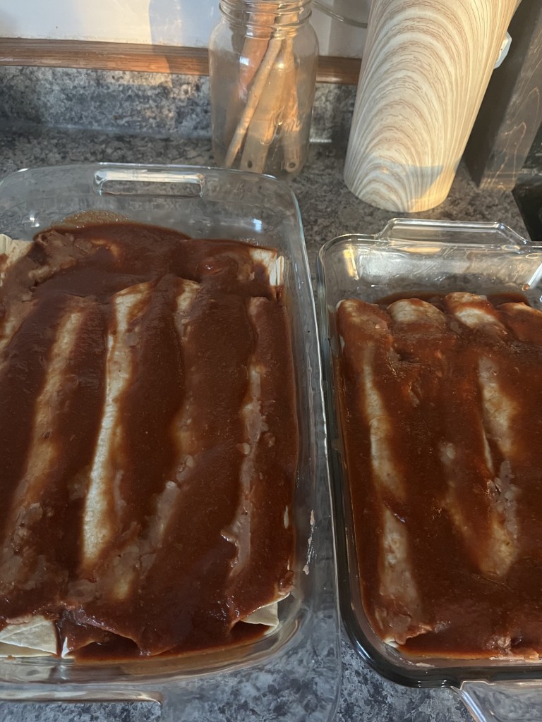 Two dishes of enchiladas with red sauce on a granite countertop. Background shows a glass jar and a striped container.