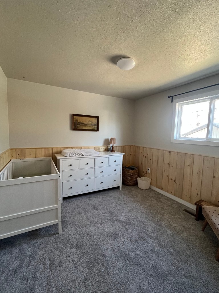A cozy nursery with a white crib, dresser, and a changing mat. Wood paneling, a window with natural light, and a framed landscape art.