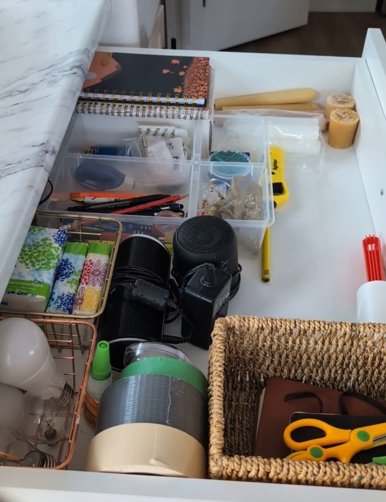 Open drawer with organized items: notebooks, tape, scissors, speakers, light bulbs, and candles. White marble surface. Neat setting.