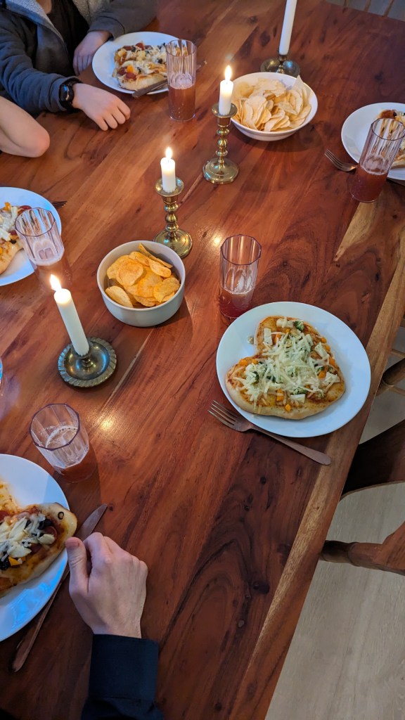 Dinner scene with people eating pizza at a wooden table, lit by candles. Plates of chips and glasses of drinks are also visible. Cozy atmosphere.