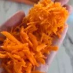 Hand holding vibrant orange grated carrots over a marble surface, conveying a fresh and textured culinary preparation.