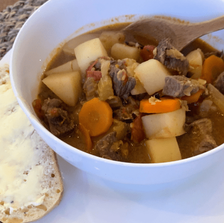 Bowl of beef stew with carrots and potatoes, stirred with a wooden spoon, next to a buttered slice of bread on a white plate.