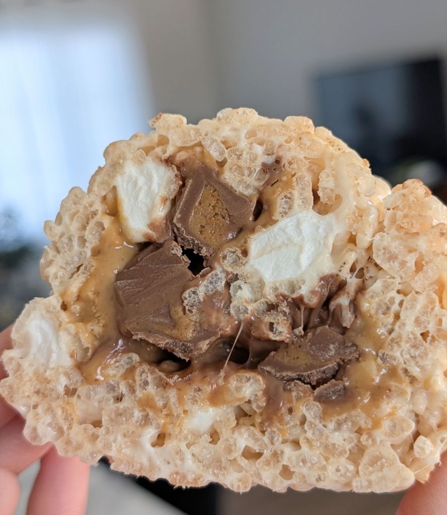 Close-up of a rice cereal treat with peanut butter, chocolate, and marshmallows. Held in hand, with a blurred indoor background.