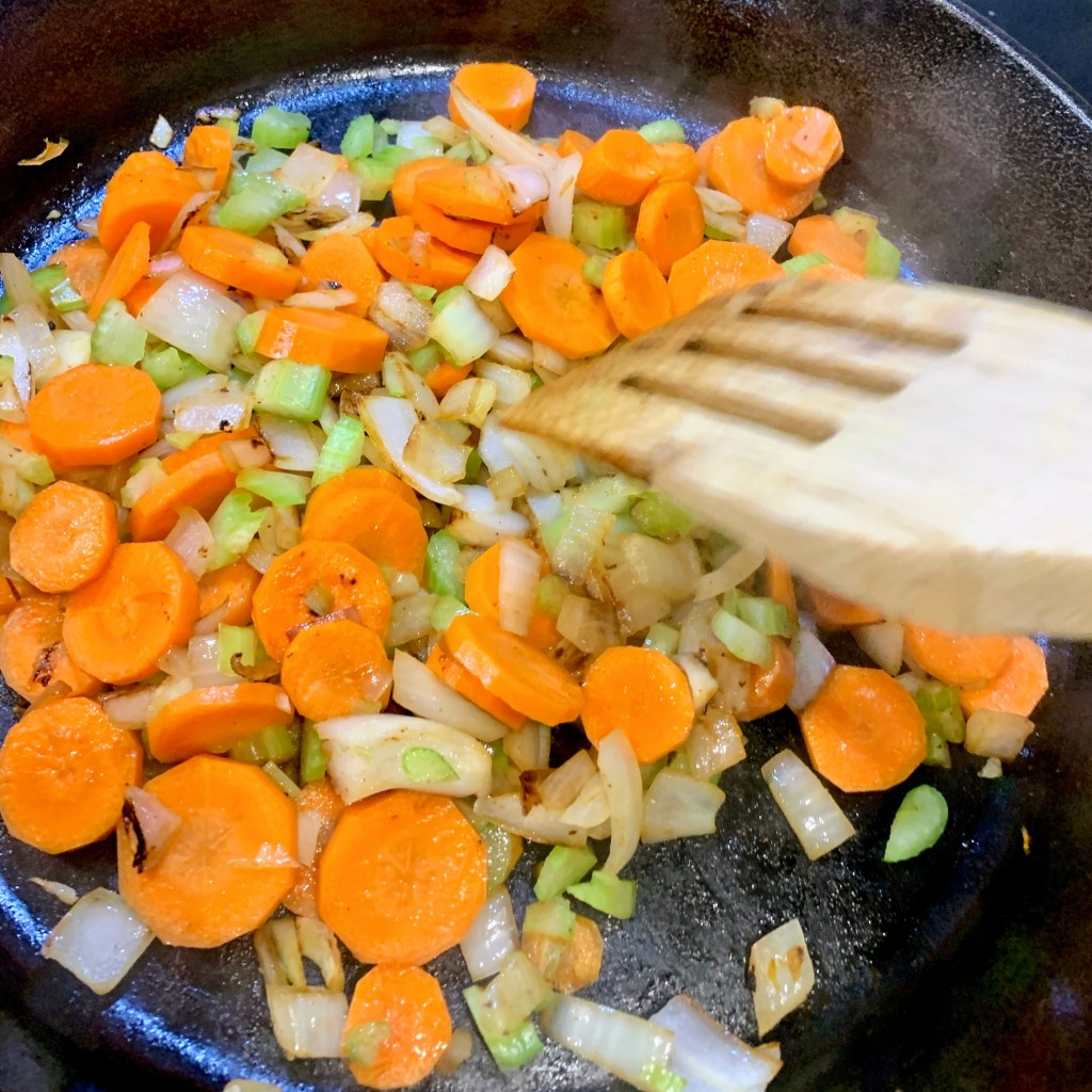Carrots, celery, and onions are being sautéed in a black pan with a wooden spatula. The colorful veggies create a vibrant contrast.