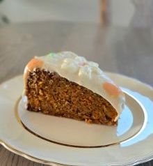 "Grandma's Carrot Cake" with white frosting and a decorative topping on a white plate. It's set on a wooden table in a bright room.