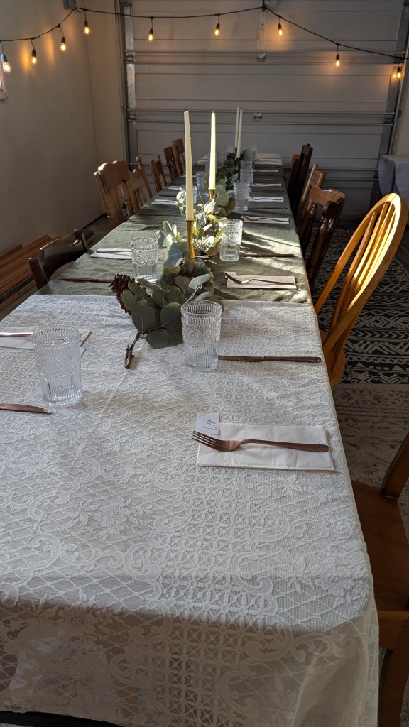 Elegant table setup with lace cloth, candles, and greenery. Clear glasses and cutlery are placed under warm string lights in a cozy room.