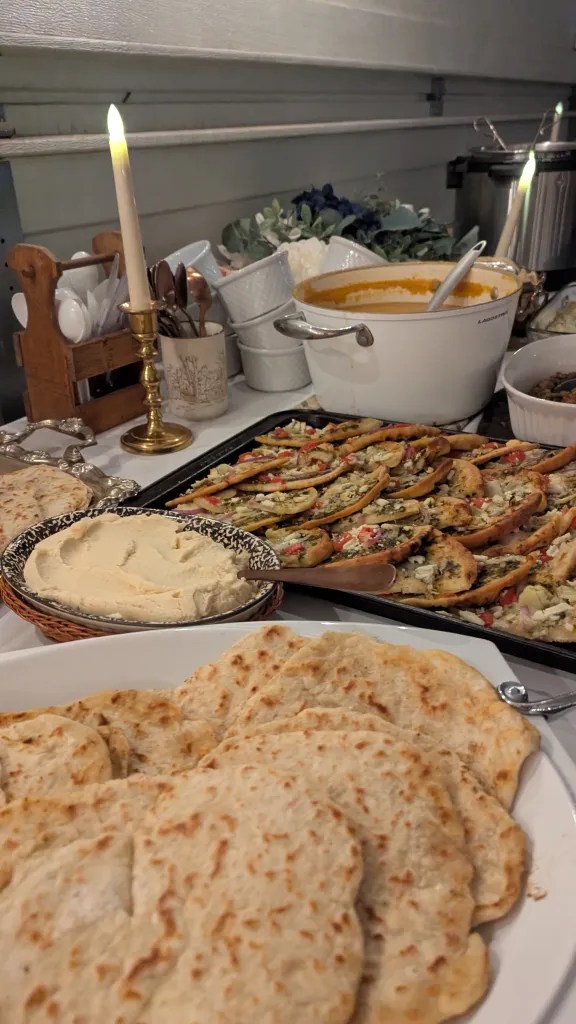 A table with flatbreads, hummus, roasted veggies on bread, soup pot, and candles. White and gold tones create a cozy, inviting setting.