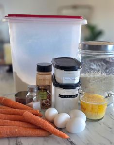 Ingredients for carrot cake are arranged: carrots, flour, sugar, eggs, butter, and spices on a marble countertop. Baking jars are labeled.