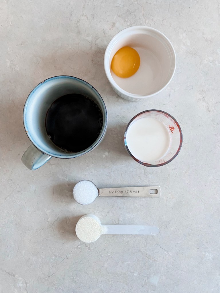 Egg yolk in a cup, black coffee, milk, sugar, and powder on a light counter. Measuring spoon reads "1/2 tbsp (7.5 ml)."