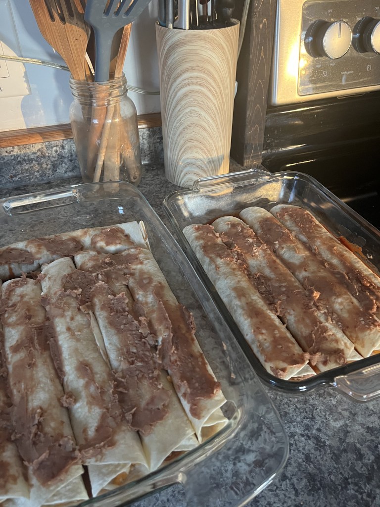 Two glass baking dishes with rolled tortillas covered in refried beans on a gray countertop. Wooden utensils and a stove in the background.