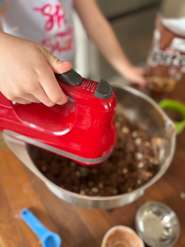 A child's hand uses a red hand mixer to blend ingredients in a metal bowl. A bag of chocolate chips and measuring cups are visible on a wooden table.