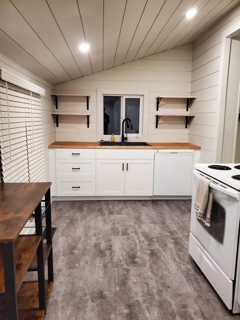 Modern kitchen with white cabinets, dark countertops, and open shelves. Gray flooring, wood accents, and a white stove with towels. Cozy ambiance.