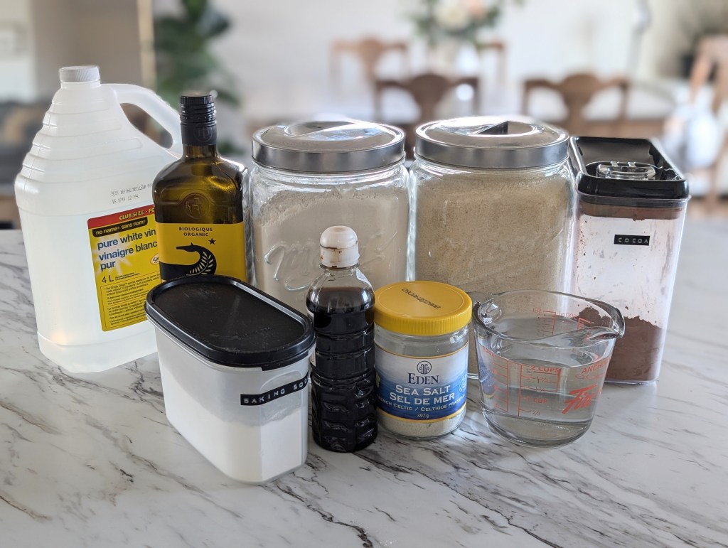 Various kitchen ingredients for ho-ho cake, on a marble countertop, including vinegar, olive oil, flour, cocoa labeled jars, and a measuring cup with water.