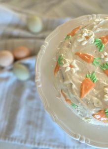 "Grandma's Carrot cake" with white frosting and orange carrot decorations on a stand. Blurred eggs in the background on a patterned cloth.