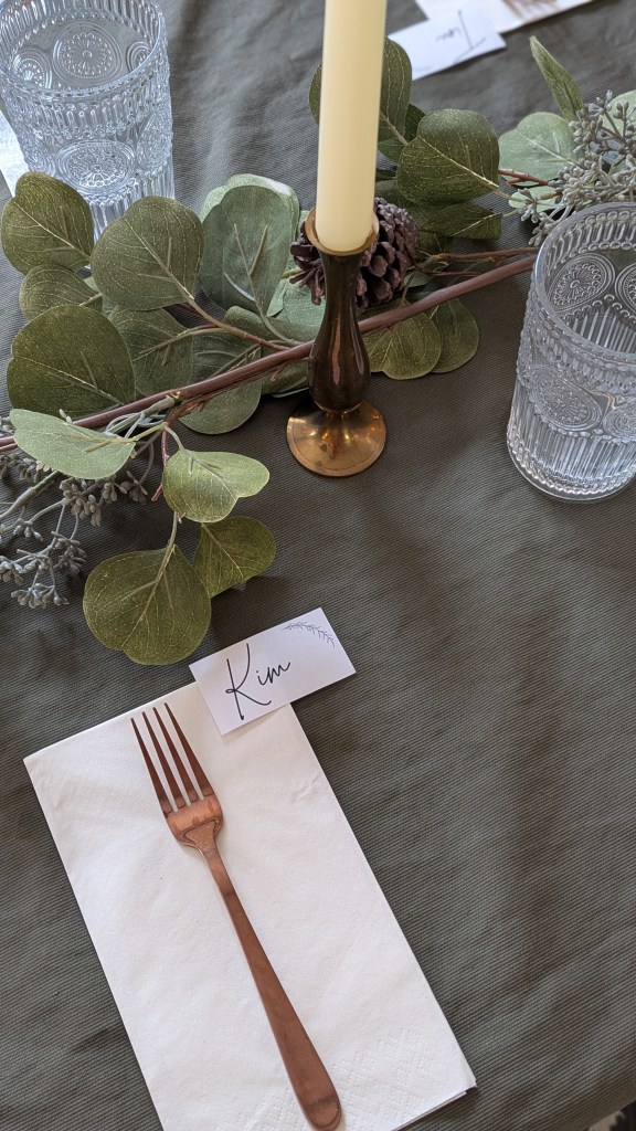 Elegant table setting with a green tablecloth, crystal glasses, featuring a centre-piece of leafy decor, and a candle. A card with "Kim" sits beside a fork on a napkin.