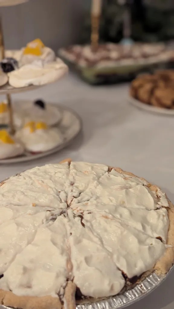 A cream-topped pie in the foreground on a table, with a cake stand and assorted desserts in the blurred background.