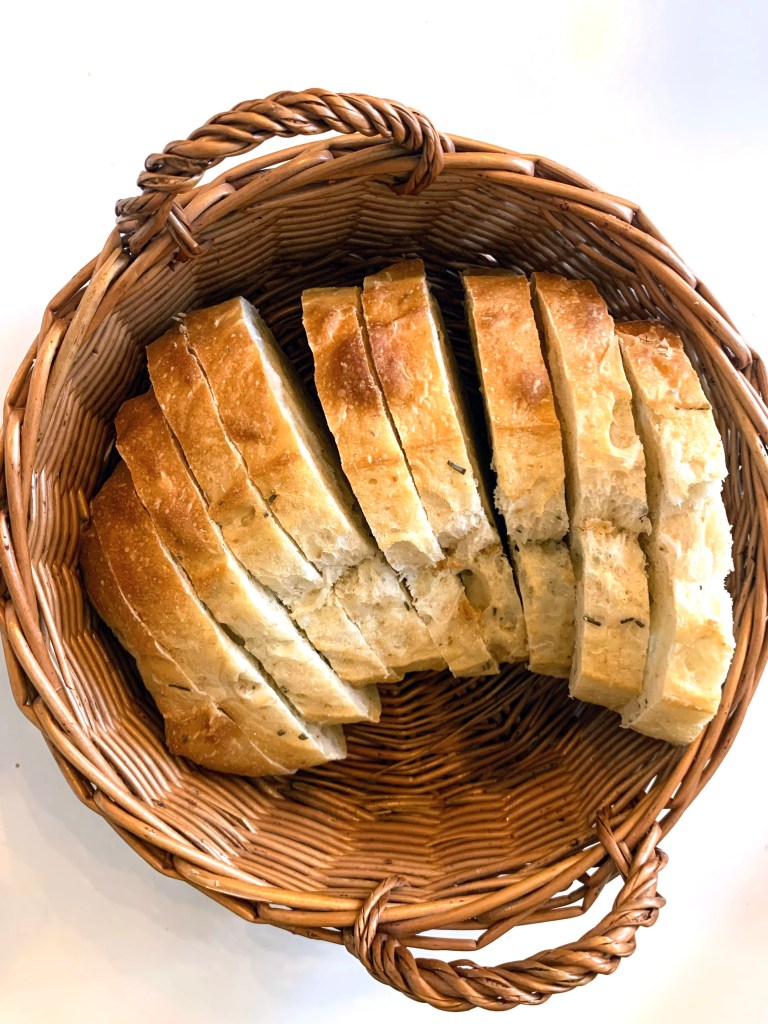 Sliced rosemary sourdough bread in a woven wicker basket, placed on a white surface. The setting is simple, with a cozy and inviting mood.