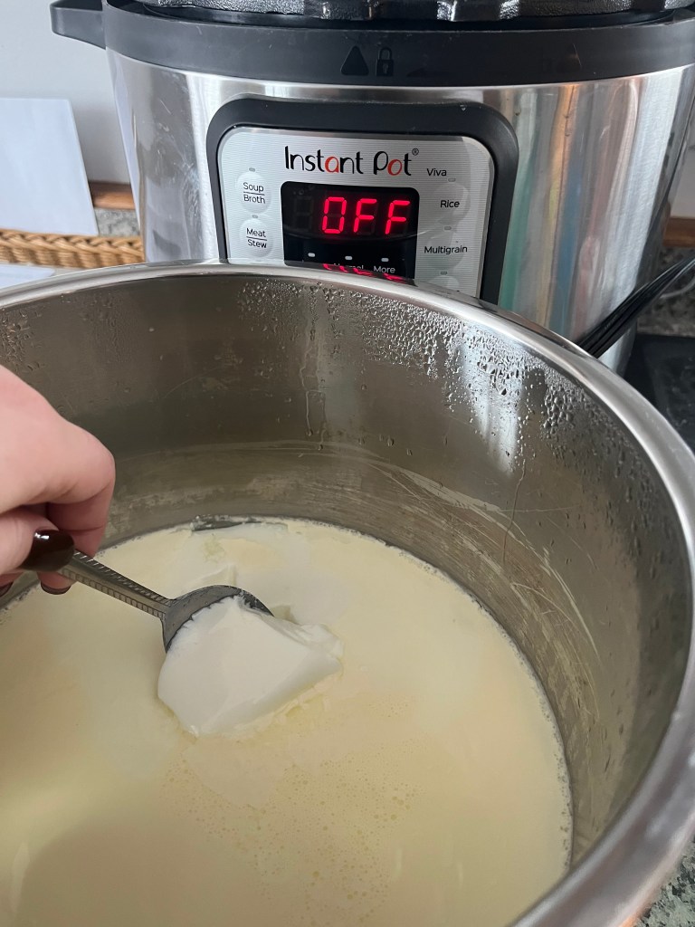 A hand holds a spoon in creamy yogurt inside an Instant Pot. The display reads "OFF." The setting is a kitchen counter.