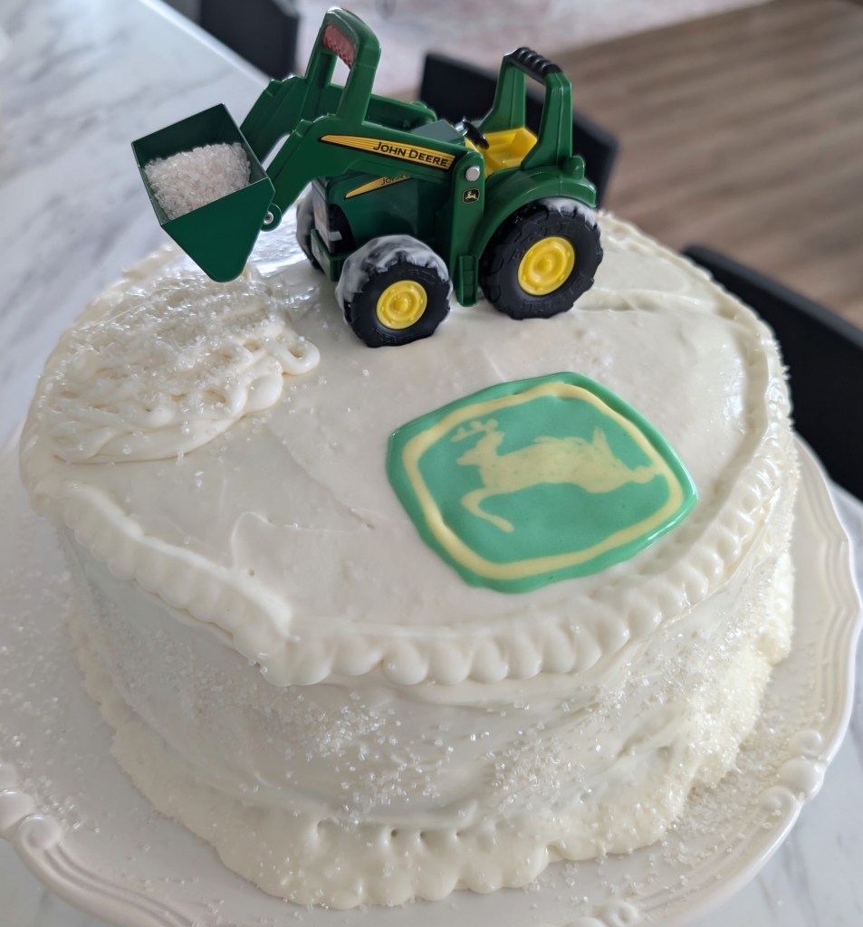 Toy tractor on white frosted cake with green logo. Tractor's scoop holds sprinkles. Cake on marble surface, casual indoor setting.