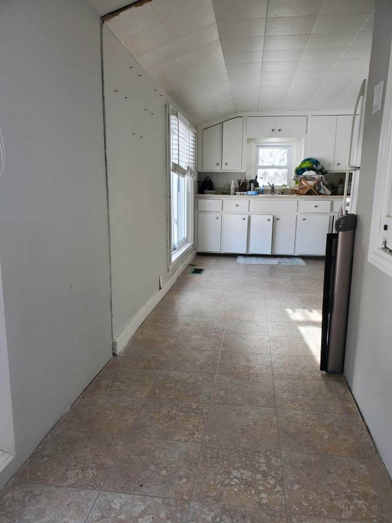 Empty kitchen with white cabinets, cluttered counter, and single window. Tiled floor and neutral walls create a simple, serene atmosphere.