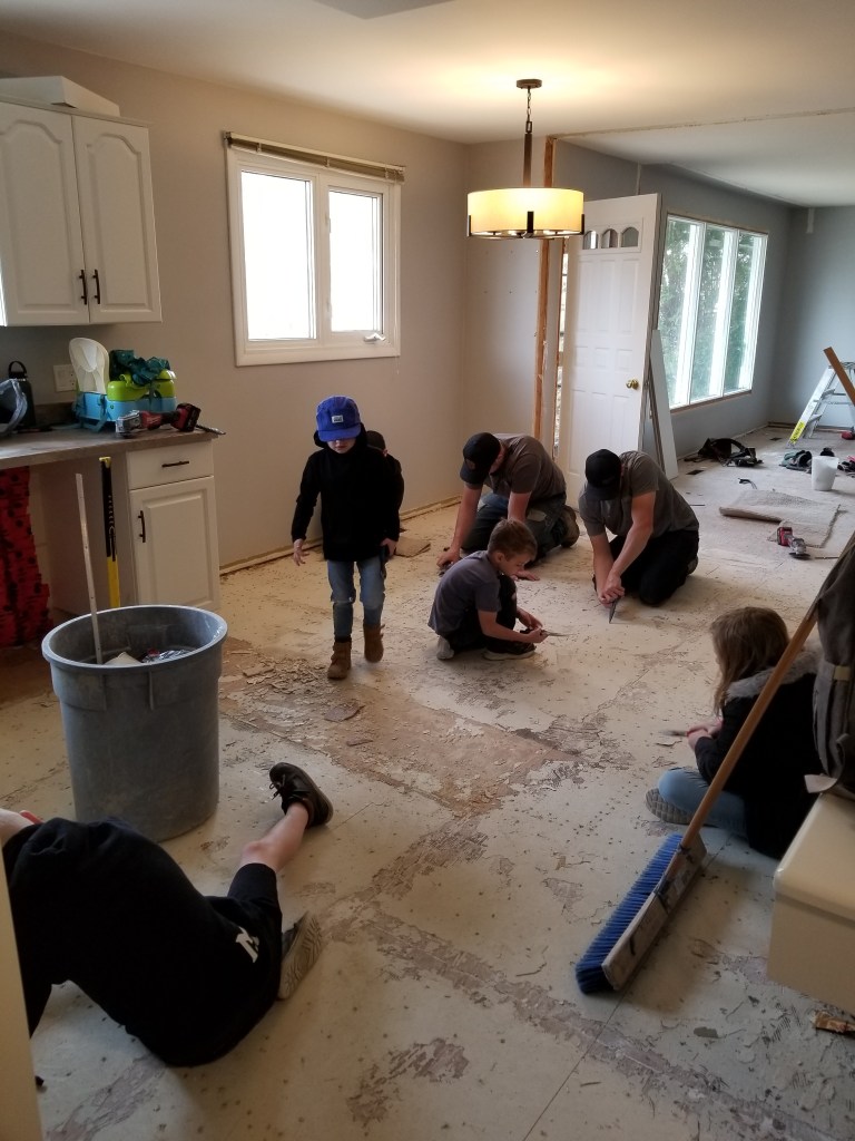 People removing floor tiles in a kitchen with white cabinets. A child wearing a blue hat helps. Bright windows and a broom visible.