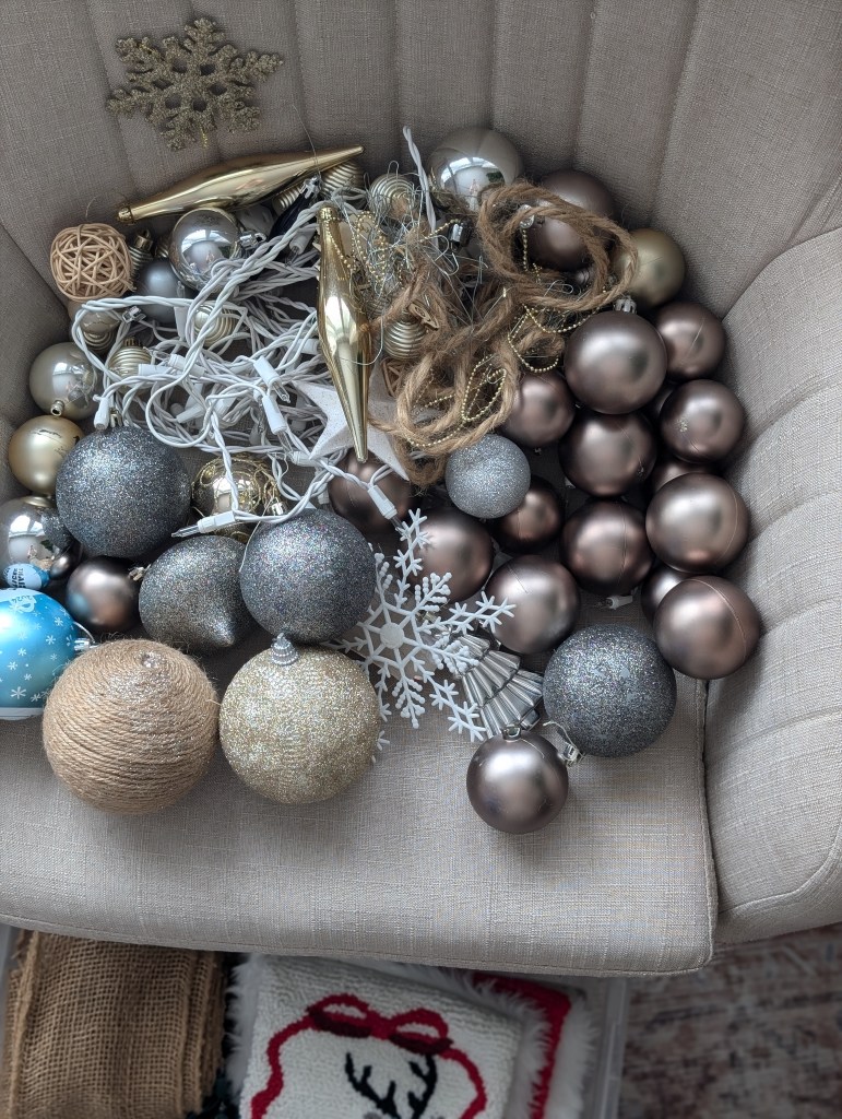Assorted Christmas ornaments on a beige chair, including gold, silver, and brown baubles, string lights, and snowflakes, evoking a festive mood.