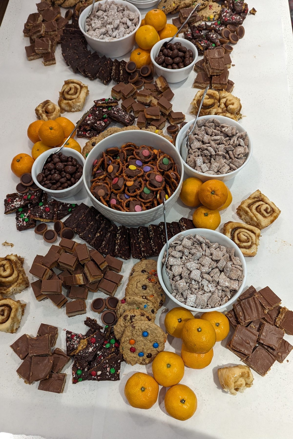 A table with assorted treats: cookies, chocolates, pretzels, and mandarins. White bowls on a white surface create a festive snack display.