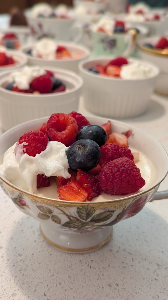 Dessert cups topped with raspberries, blueberries, and cream on a countertop. Floral-patterned cup in foreground with a bright, inviting mood.