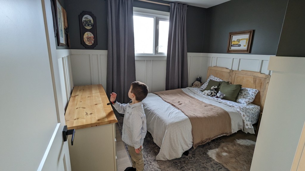Child in a cozy bedroom looks at framed wall art. Room features a wooden headboard, neutral bedding, and plush toys. Gray curtains frame a window.