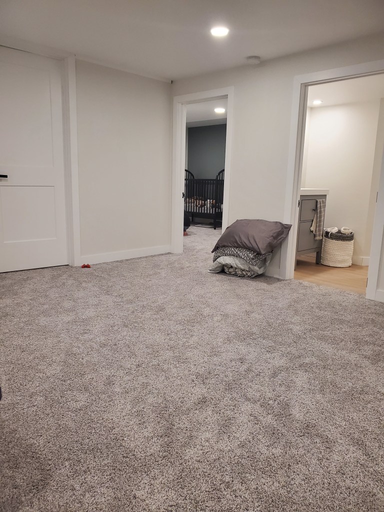 Carpeted room with two open doors; one shows a crib in a dimly lit nursery, the other leads to a laundry basket. Neutral tones and a pillow stack.