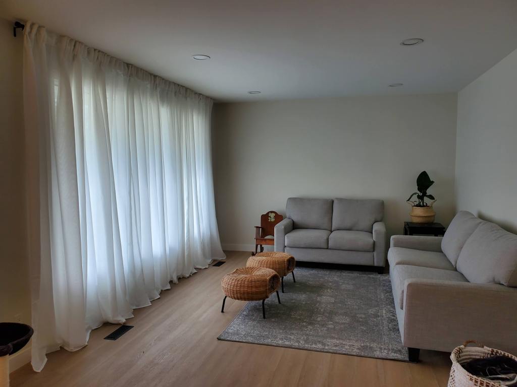 Minimalist living room with two gray sofas, wicker stools, and a potted plant. Sheer white curtains cover large windows, creating a serene vibe.